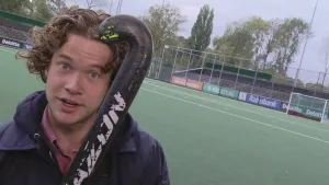 Foto van presentator, je ziet met name zijn hoofd, met een hockeystick, op de achtergrond zie je een hockeyveld.