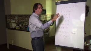 Foto van presentator die staat voor een groot hangend vel papier waarop aantal wiskundige sommen staan geschreven.