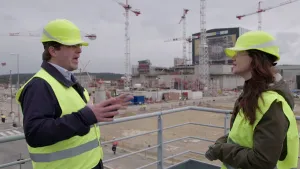 Foto van man en vrouw in beschermende werkkleding en helmen op die staan op een stijger op een groot terrein waarop gebouwd wordt.