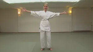 Foto van presentator in sportkleding van Aikido die in sportzaaltje een staande oefening doet.