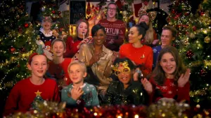 Foto van groep kinderen en volwasenen met Edsilla Rombley, in Kerstskleding, die Kerstliedjes zingen, er staan Kerstbomen omheen.