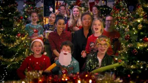 Foto van groep kinderen en volwasenen met René Froger, in Kerstskleding, die Kerstliedjes zingen, er staan veel Kerstbomen omheen.