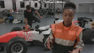 Foto van presentator in grote garageruimte waarin een raceauto van Formule 2 staat, erachter is een man bezig aan die auto.