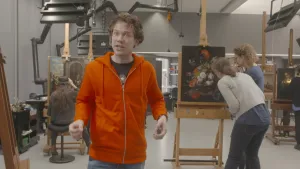 Foto van grote werkruimte waarin schilderijen worden gerestaureerd, een vrouw is aan een schilderij bezig, presentator staat ervoor.