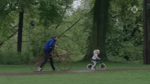Foto van een man die loopt naast een ouderwetse fiets, voor hem uit fiets jong meisje op een kinderfietsje.