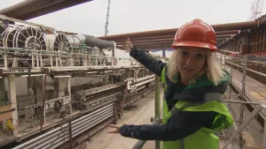 Foto van presentator in werkkleding en helm op die buiten staat bij een tunnelboor, groot langwerpig apparaat.