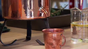 Foto van soort koperen vat met tuitje waaruit doorzichtige vloeistof komt en terecht komt in een koperen beker.