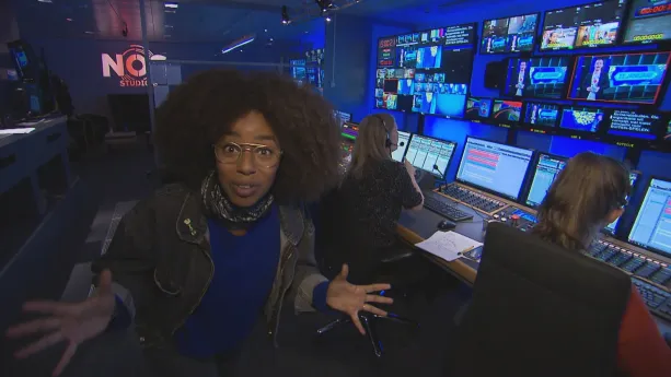 Foto van presentator die staat in de studio van het Jeugdjournaal, je ziet veel beeldschermen en computers met mensen erachter.