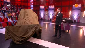 Foto van de Studio Snugger waar op het podium de presentator staat bij een groot voorwerp waarover een grote doek ligt, niet te zien wat het is.