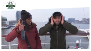 Foto van man en vrouw die bij de Maas in de stad Rotterdam staan, ze kijken allebei alsof ze enorm lawaai horen.