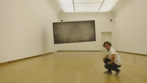 Foto van een museumzaal waarin een grijze rechthoek hangt, erbij zit presentator gehurkt op de grond.