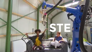 Foto van een fabrieksruimte waarin een enorme robotarmachtige machine te zien is en presentator die een 3D-geprinte klokhuis vasthoudt.