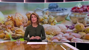 Foto van presentator aan de desk in de studio met achter haar grote foto van allerlei voedingsproducten zoals broodjes en eieren.
