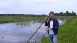 Foto van presentator met lange stok van gereedschap voor turfwinning die staat aan de over van een natuurlijk water met grasland.