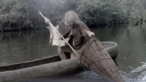 Foto van man die op soort roeiboot zit en bezig is met een fuik, op het water, hij is gekleed zoals heel lang geleden.