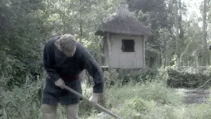 Foto van man die staat in het bos met een stok van gereedschap in het bos, er staat een klein houten hutje.