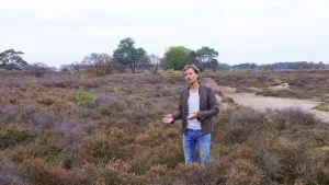 Foto van presentator die staat op de heide nabij een pad, heidegebied met wat bomen.