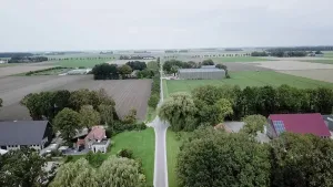 Foto van een smalle weg door weilandengebied met boerderijen en loodsen/schuren erlangs.