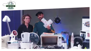 Foto van man en vrouw die staan voor een tafel met elektrische huishoudelijke apparaten erop zoals een magnetron, de man houdt twee vliegtickets vast.