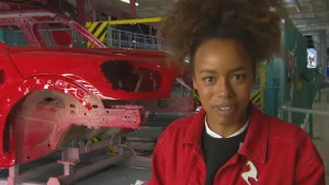 foto van presentator die staat in de autofabriek, je ziet een deel van een auto op de lopende band.