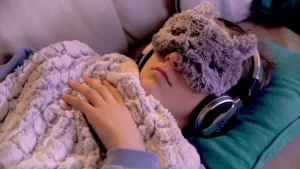 Foto van jongetje dat slaapt op de bank, dekentje om, lapje over de ogen, heeft oortelefoon op.