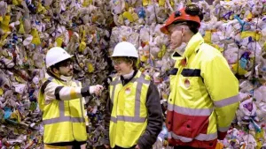 Foto van twee kinderen en man in werkkleding bij een muur gemaakt van allemaal plastic afval.