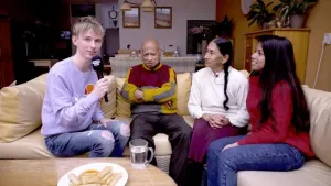 Foto van aantal mensen, jong en volwassen, op grote bank in huiskamer, één van hen heeft een microfoon in de handen.