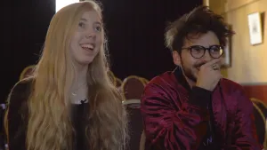 Foto van een man en een lachend meisje die naast elkaar in een kamer zitten en ergens naartoe kijken.