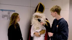 Foto van een Sinterklaas die staat bij een jongen met microfoon die hem interviewt en een meisje.