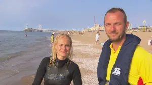 Foto van man en vrouw in zwem-/reddingspak die staan op het strand van Scheveningen met mooi weer.