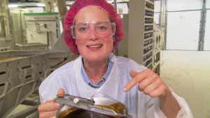 Foto van presentator in beschermende werkkleding in fabriek met soort bak met bruine vloeistof erin voor het maken van drop.