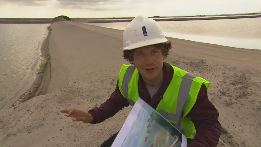 Foto van presentator in werkkleding met een kaart in de handen die staat bij een lange pas aangelegde zanddijk in het water.