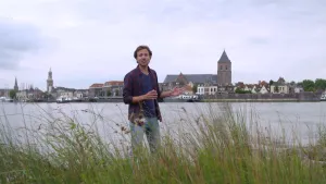 Foto van de skyline van een stad in Nederland aan overkant van een rivier, op de oever aan deze kant staat presentator.