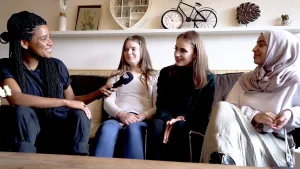 Foto van presentator met microfoon die twee meisjes interviewt die op de bank in woonkamer zitten.