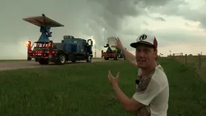 Foto van presentator die staat voor een weg waarop aantal wagens met meetapparatuur rijden, bewolking van bui, storm.