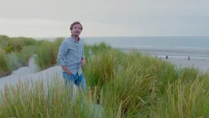 Foto van presentator die op een duin in duingebied staat, op de achtergrond de zee.