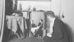 Oude foto, zwart/wit, van een man die in kamer zit, in een kast zitten paar onderduikers, ten tijde van de Tweede Wereldoorlog.