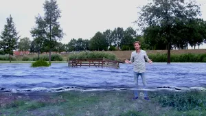 Foto van presentator die in het water staat van een rivier met hoog water, middenin het water staat een houten hek.