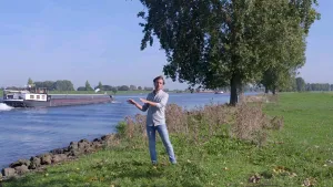 Foto van presentator die staat op de oever van een rivier waarover een vrachtboot vaart.