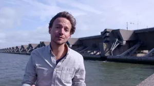 Foto van presentator die staat bij water met één van de Deltawerken, een lange waterkering.