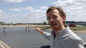 Foto van presentator die staat bij een autobrug over breed water, rivier.