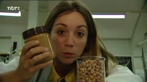 Foto van presentator in laboratoriumachtige ruimte, je ziet haar gezicht, die een pot pindakaas vasthoudt en voor haar staat glazen pot met pinda's.