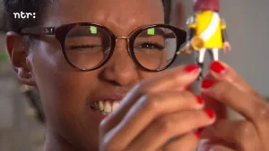 Foto van presentator, portret, met die door een bril kijkt naar een klein plastic poppetje.