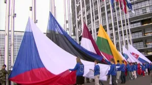 Foto van een reeks vlaggen beneden aan de stok bij een modern gebouw, er staan mensen bij die ze gaan hijsen.