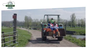 Foto van een tractor met aantal mensen erop die over een weggetje rijdt in weidelandschap.