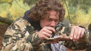 Foto van man in camouflagekleding die met een vergrootglas kijkt naar een stuk hout in de natuur.
