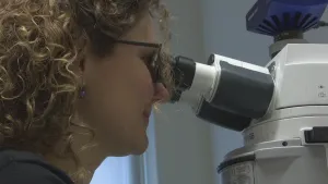 Foto van vrouw, je ziet haar hoofd, die kijkt in een groot soort microscoop, je ziet daar een deel van.
