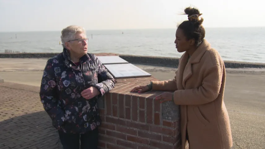 Foto van oude vrouw en presentator die staan bij een gedenkteken, op de achtergrond de zee, over de Tweede Wereldoorlog.