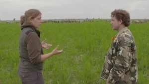 Foto van man en vrouw die staan op grote grasvlakte in een natuurgebied, op de achtergrond kudde vossen te zien.
