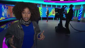 Foto ven presentator die staat in de studio van het Jeugdjournaal, je ziet een camera met cameraman, afbeelding op de achtergrond.